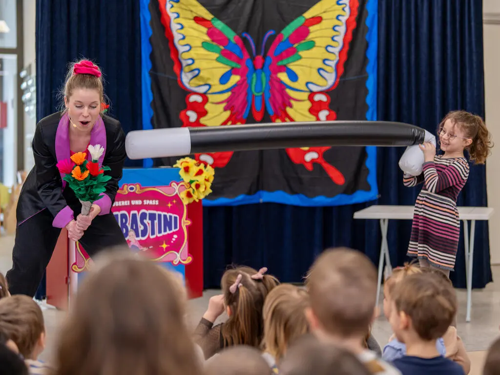 Kinderzauberin Bombastini und ein Mädchen halten gemeinsam einen Riesenzauberstab bei der Mitmach-Zaubershow vor Schulpublikum