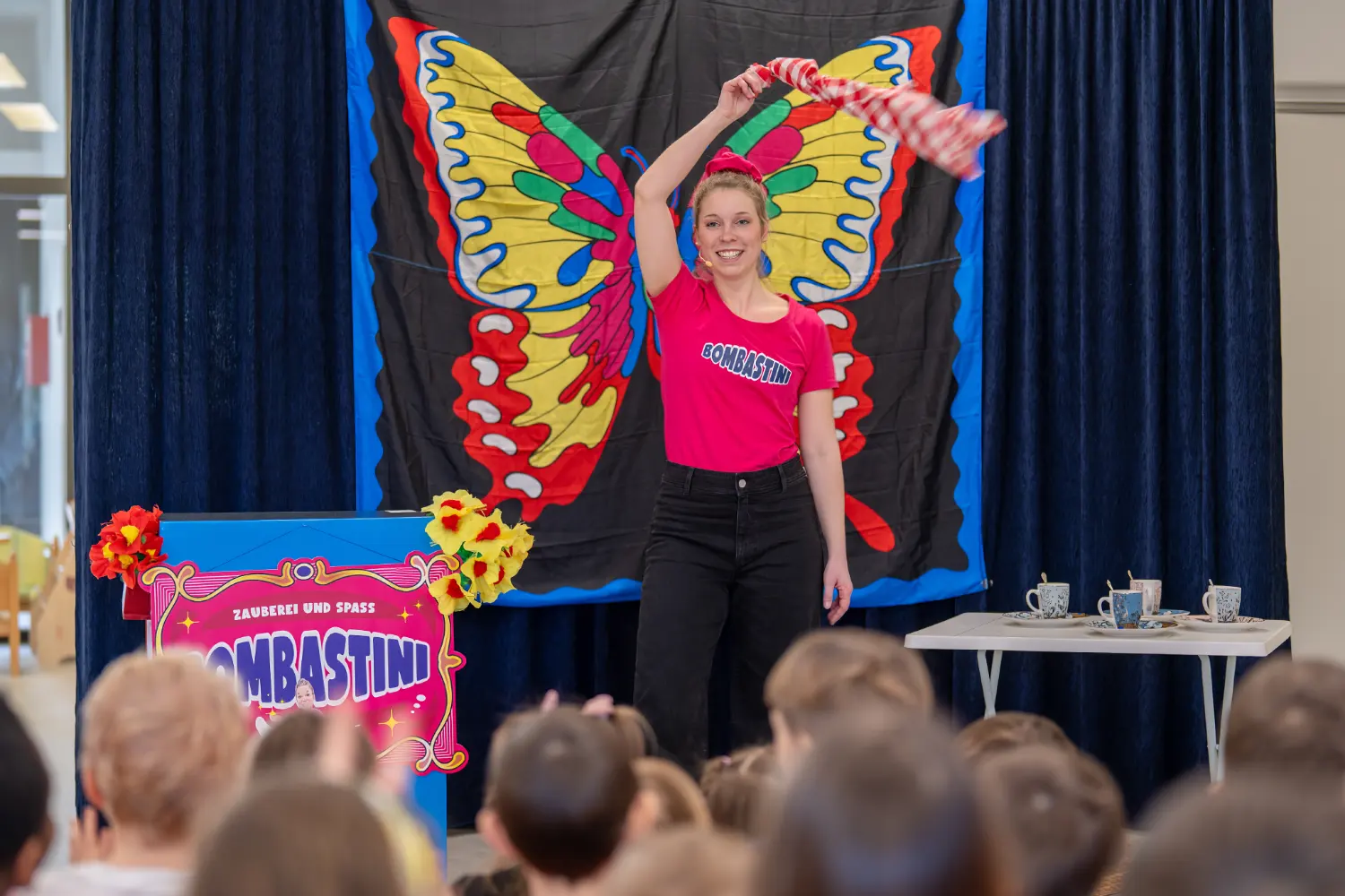 Kinderzauberin Bombastini im pinken Shirt schwenkt Zaubertuch vor großem Schmetterling-Bühnenbild vor begeisterten Kindern